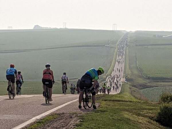 A large number of cyclists going down then up a hill in the rural distance.