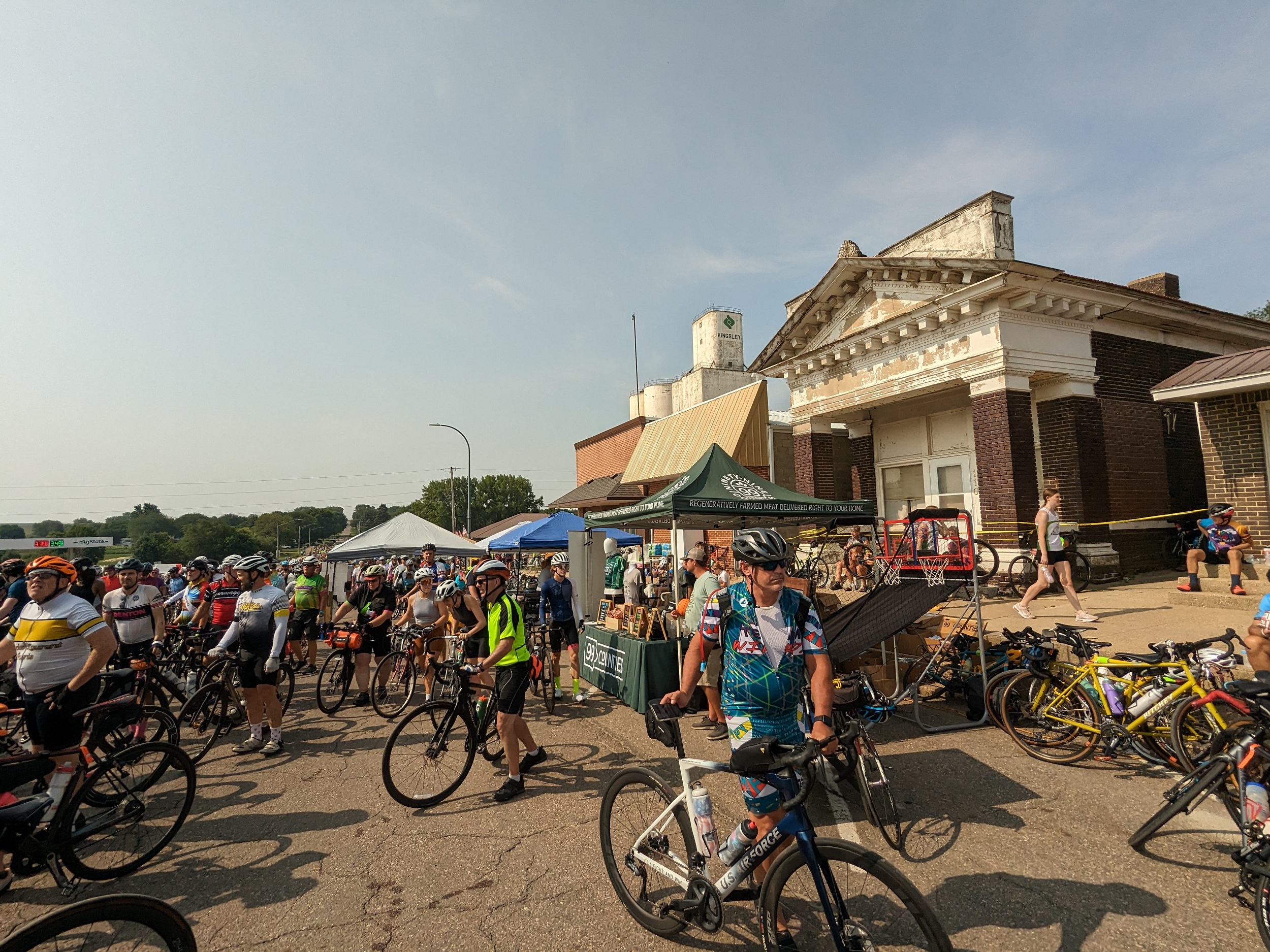 Crowd of walking cyclists with a classical revival one story and white grain elevator.
