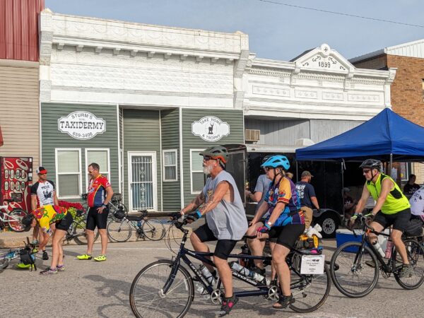 A couple on a tandem bike riding past two late 19th century one story storefronts