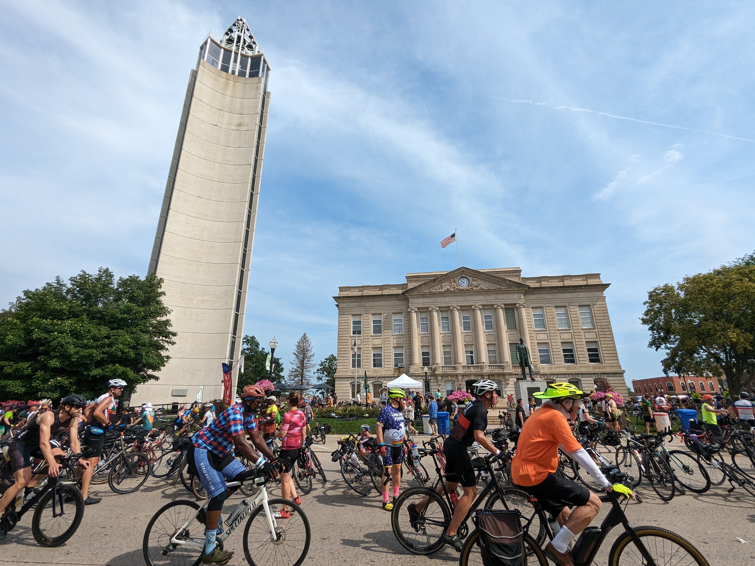 Many cyclists riding past a Classical Revival cout house and a Mid-century Modern clock tower.