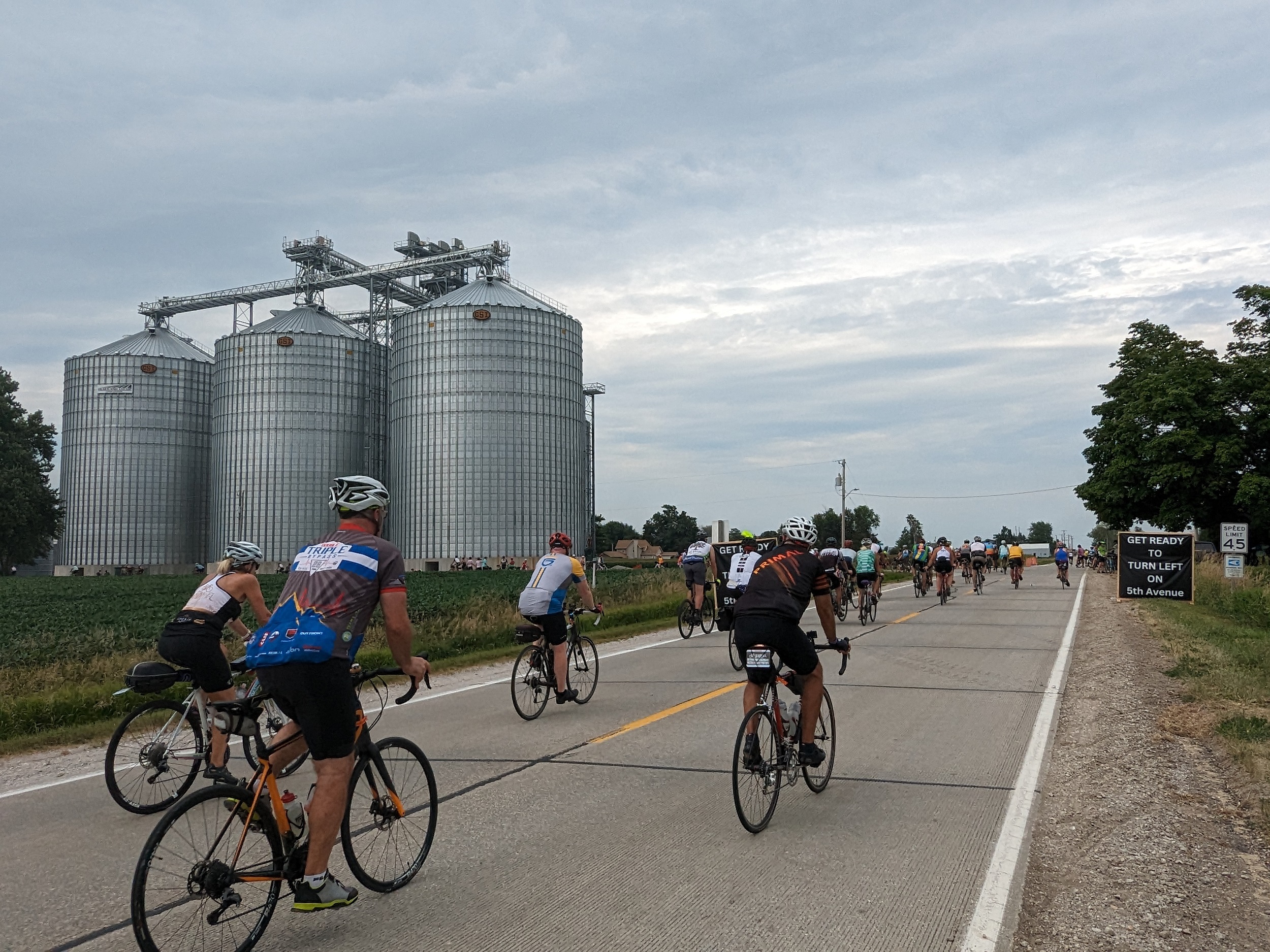 Cyclists on a state highway with clean metal grain elevators to the left.