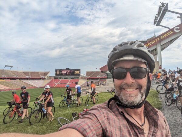 Me with many cyclists walking their bikes through a grass patch inside Jack Trice football stadium.