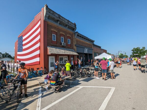 A set of three late 19th century brick storefronts, nearest with a large American flag on its long corner side and cyclists all around.