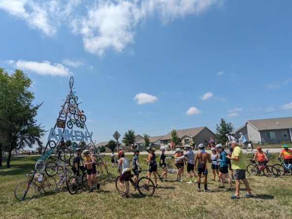 Numerous cyclists stand around a metal pyramid structure with attached bikes while taking pictures.