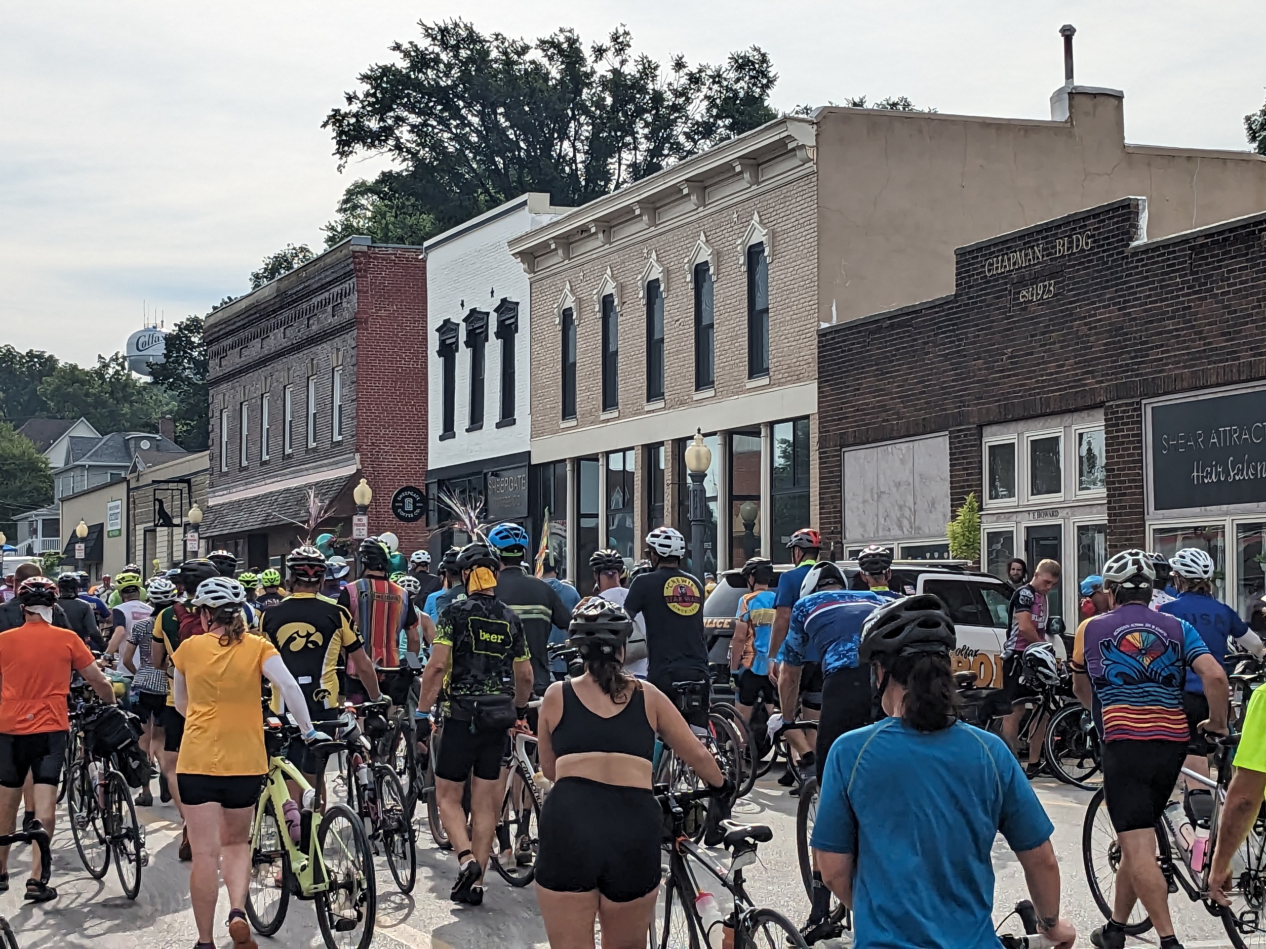 A crowd of cyclists walking their bikes through a brick late 19th century downtown of two story storefronts.