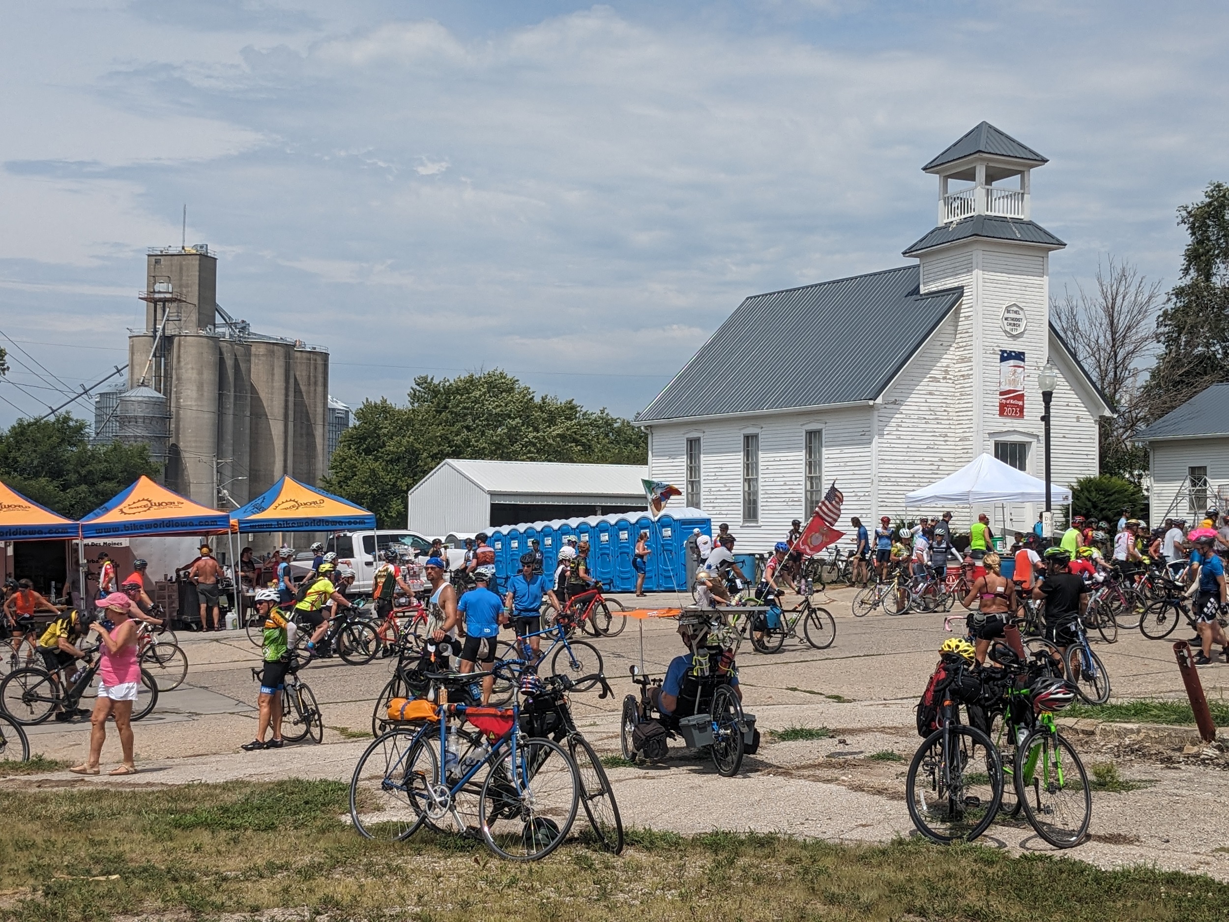 Numerous cyclists with a white wood two story church in the fore and grain elevator in the background.