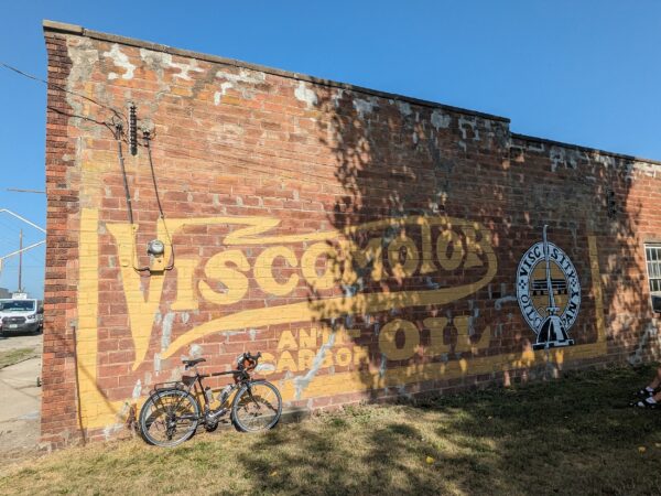 A tour bike laeninf on a outside brick wall with a touched up yellow Viscomotor and Oil ghost sign.