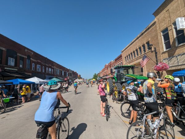 Numerous cyclist riding and standing on a main street with an uninterrupted canyon of late 19th Century buildings