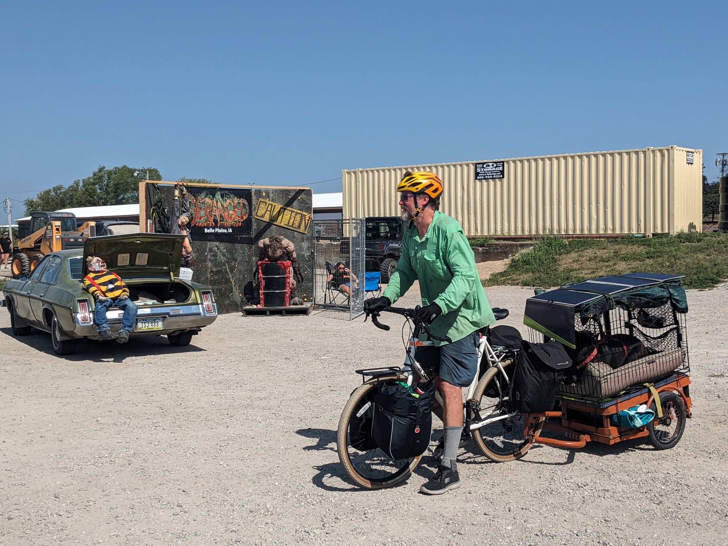 A cyclist towing a trailer holding a dog standing in front of a mock horror scene roadside attraction.