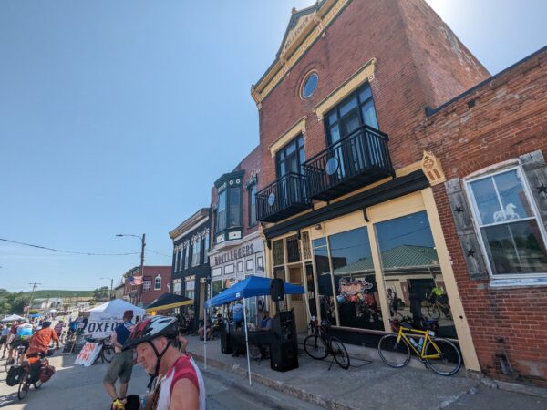 Cyclist on the left riding out of town into the distance with a small set of late 19th Century storefront and flats to the right.