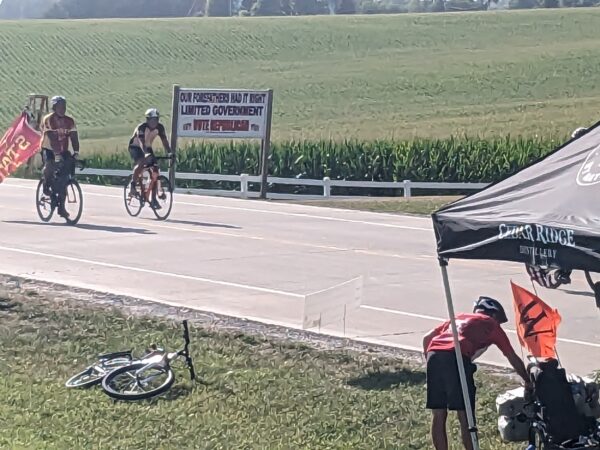 A closeup picture of two cyclist on a state road passing a pro Republican limited government sign in front of federally supported corn fields.