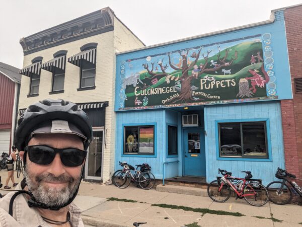 Me facing the camera with late 19th Century buildings behind, one with a colorful mural that reads Eulenspiegel Puppets