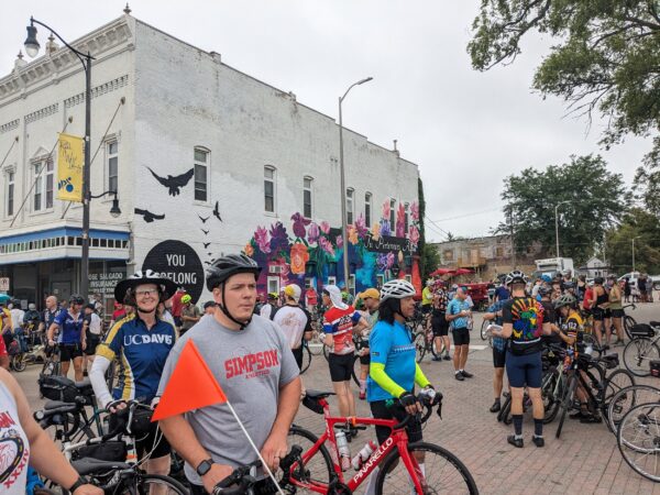 Numerous cyclists of different shapes and sizes on the street with a mural on he side of a corner building behind.