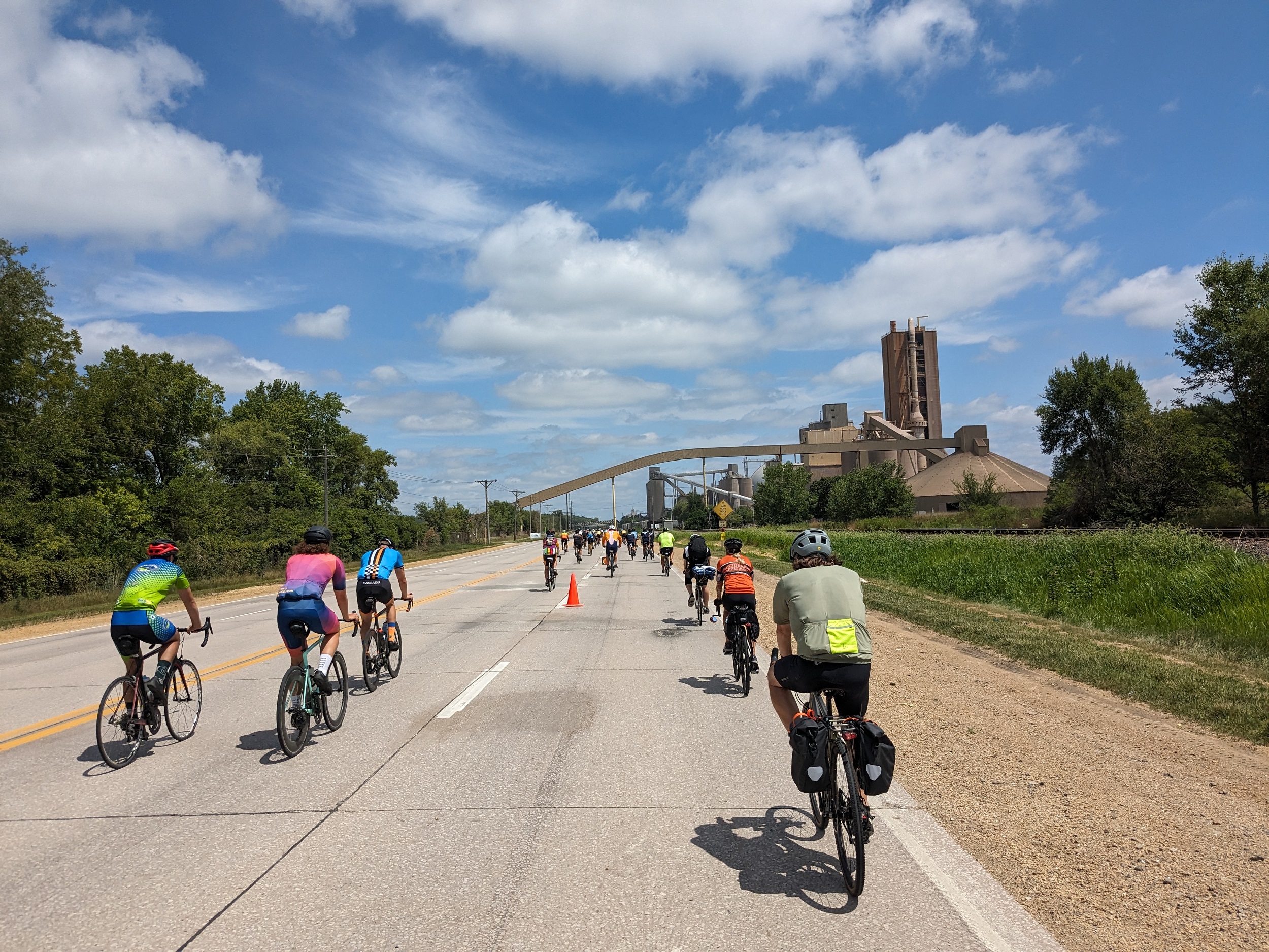 Action shot from the bike with numerous cyclists no the road riding towards an industrial facility that has a conveyor over the road.