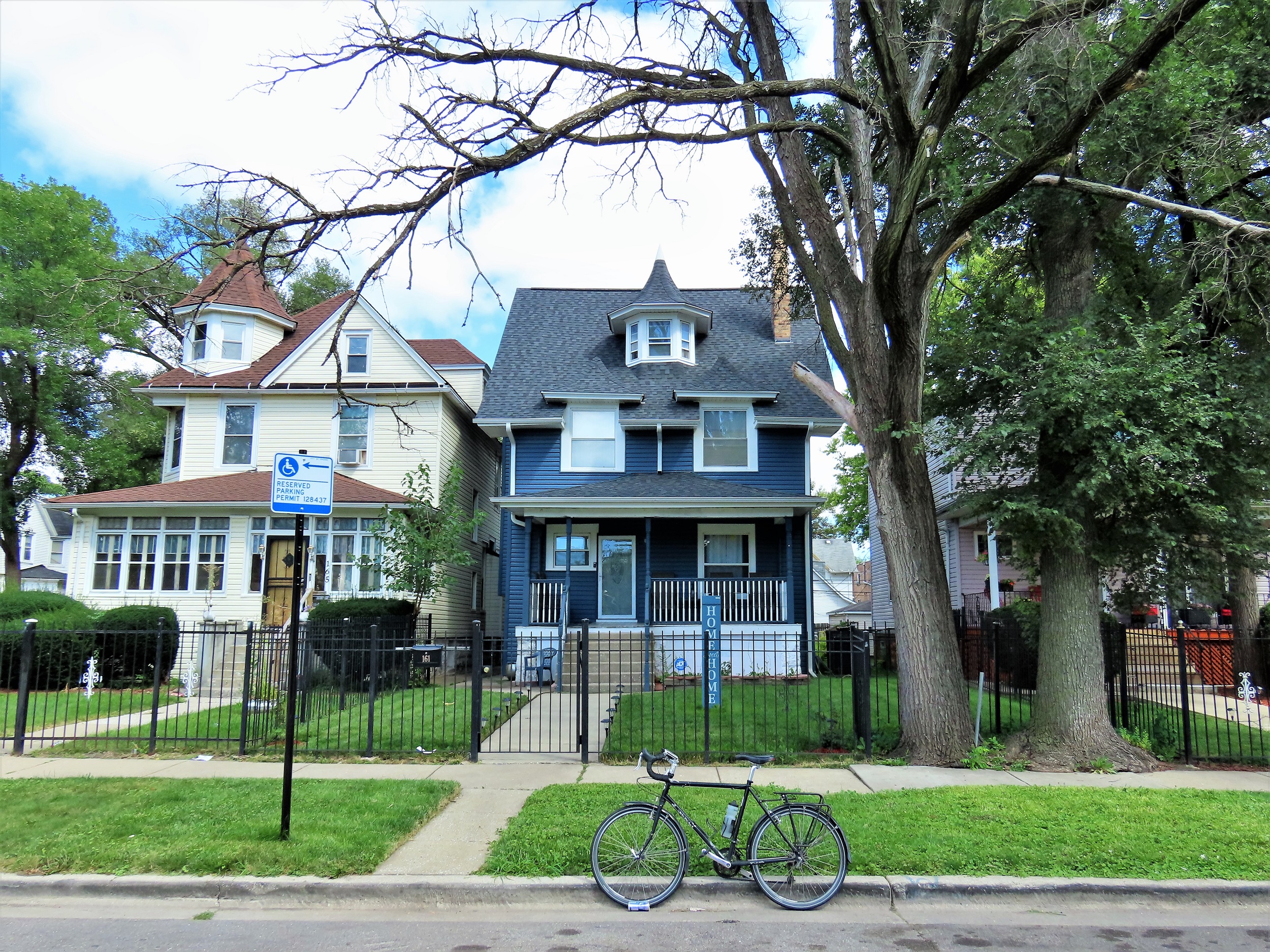 A tour bike standing at front of a three story dark blue and white trim 1890s wood residence with a small round witch hatted roof dormer in between two square witch hatted second floor windows.