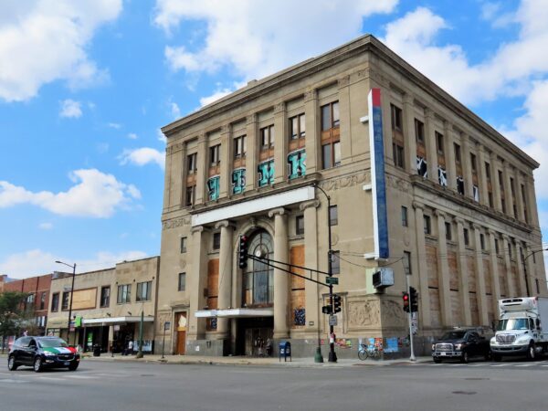 Across a large intersection a tour bike leans on the base of a five story limestone Classical Revival corner vacant former bank