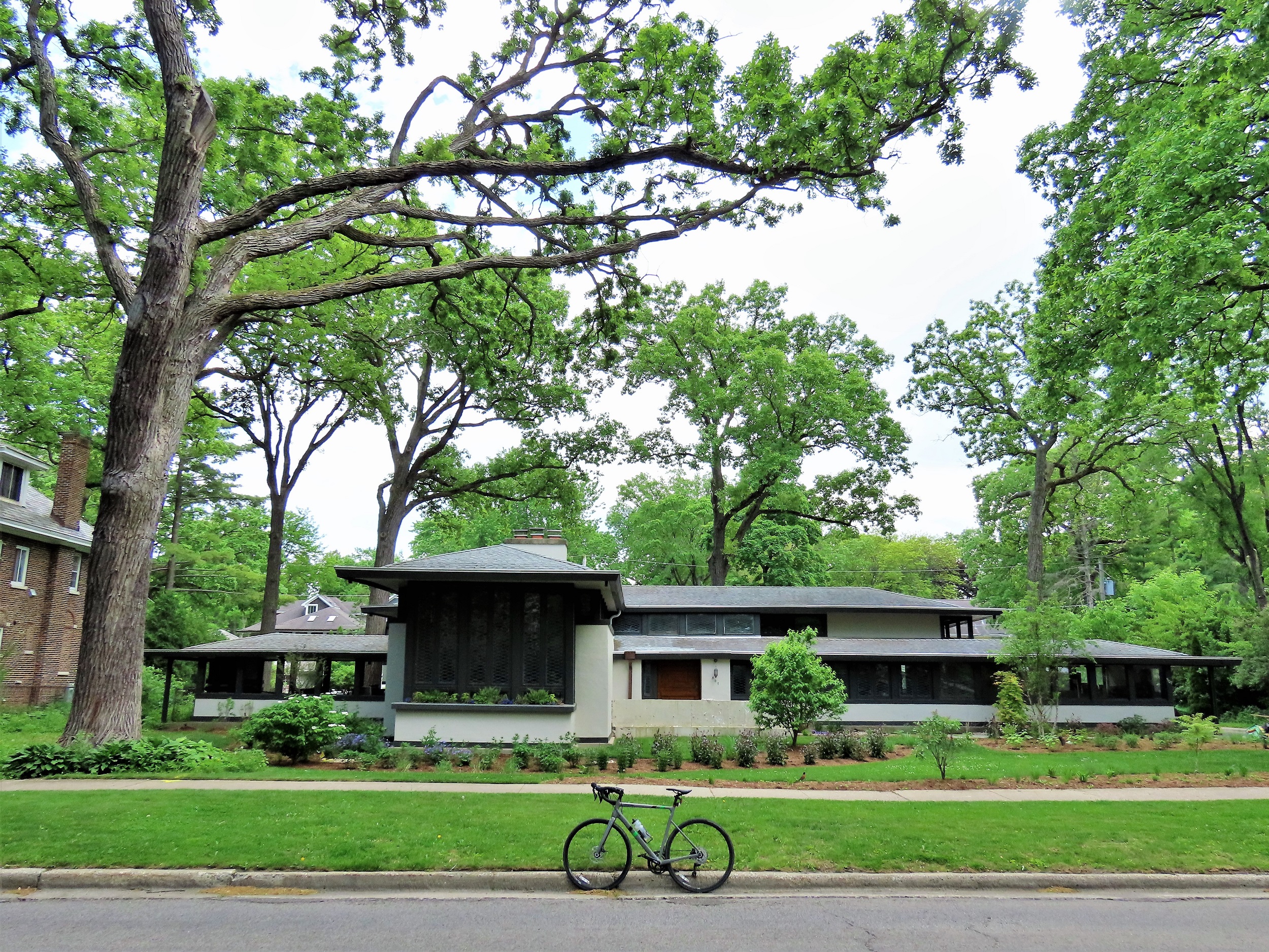 A tour bike standing at front of a two story horizontally wide low hipped roofed Prairie School style single family home