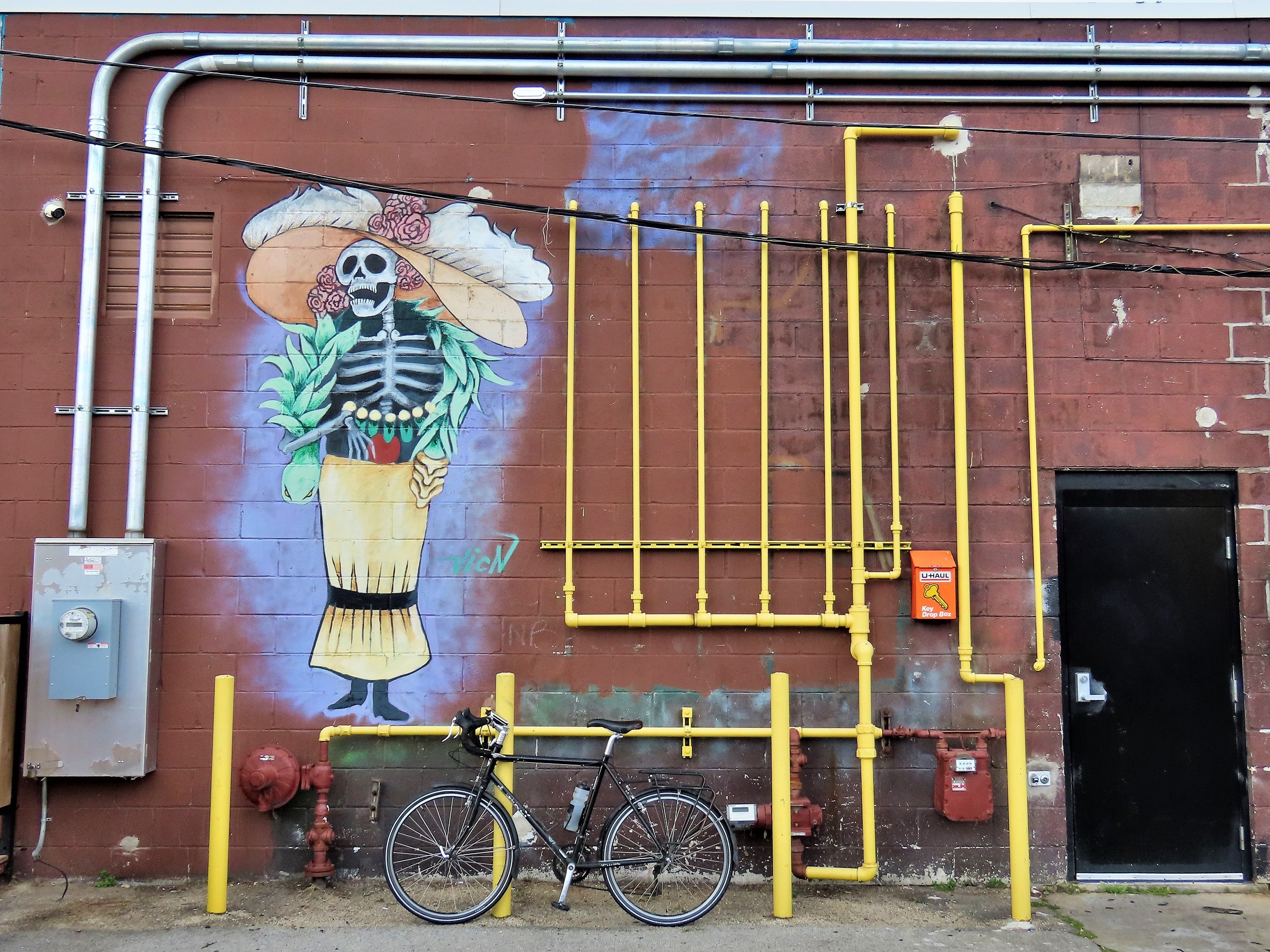 A tour bike leaning on yellow painted posts in front of a wall covered in yellow painted and unpainted pipes with a Mexican style female skeleton with grand wide hat among the pipes.