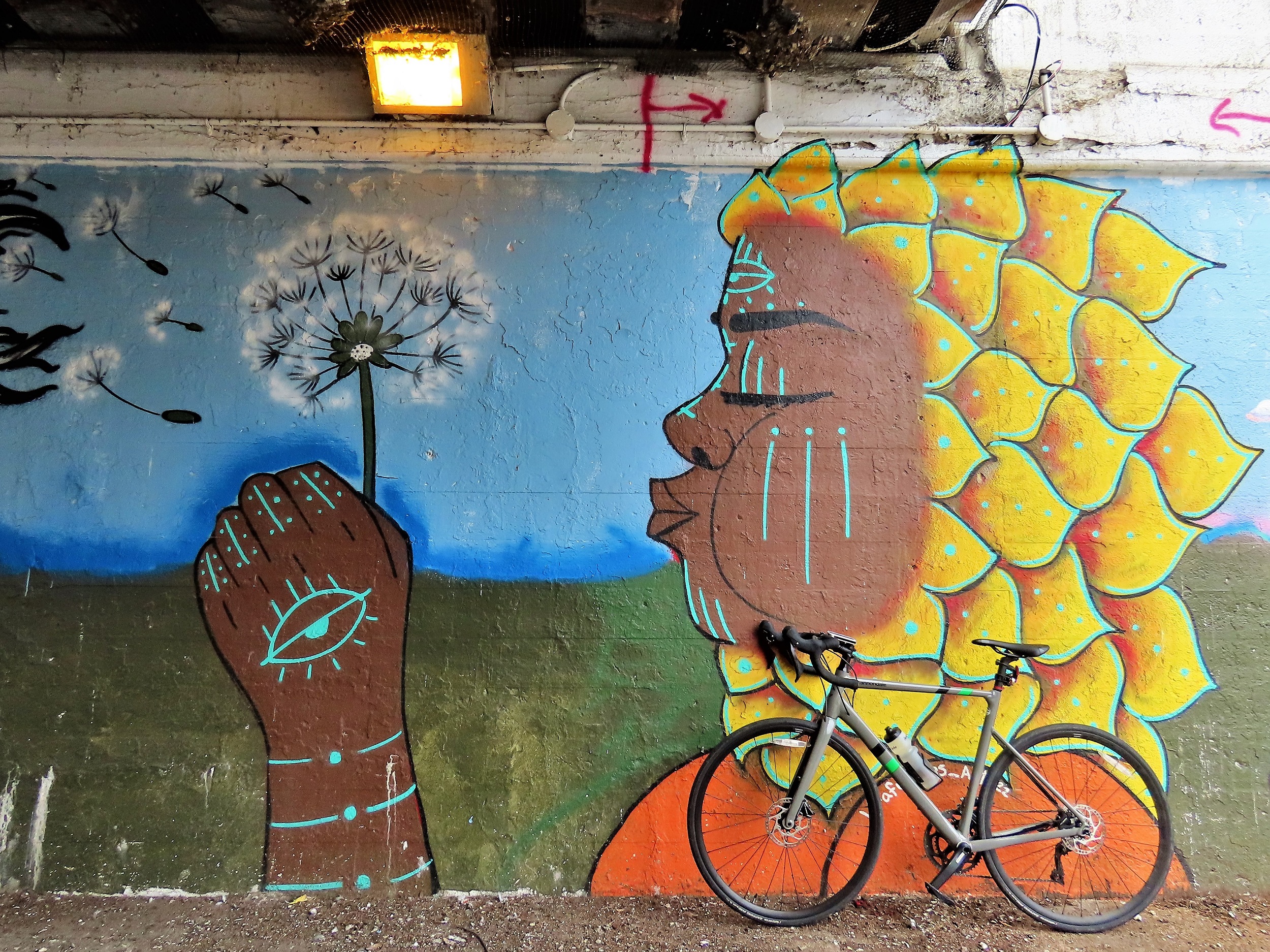 A tour bike leaning at the base of an underpass mural depicting a sunflower-shaped-hair brown skinned girl with her eyes closed mouth set to blow into the dandelion she holds in her hand.
