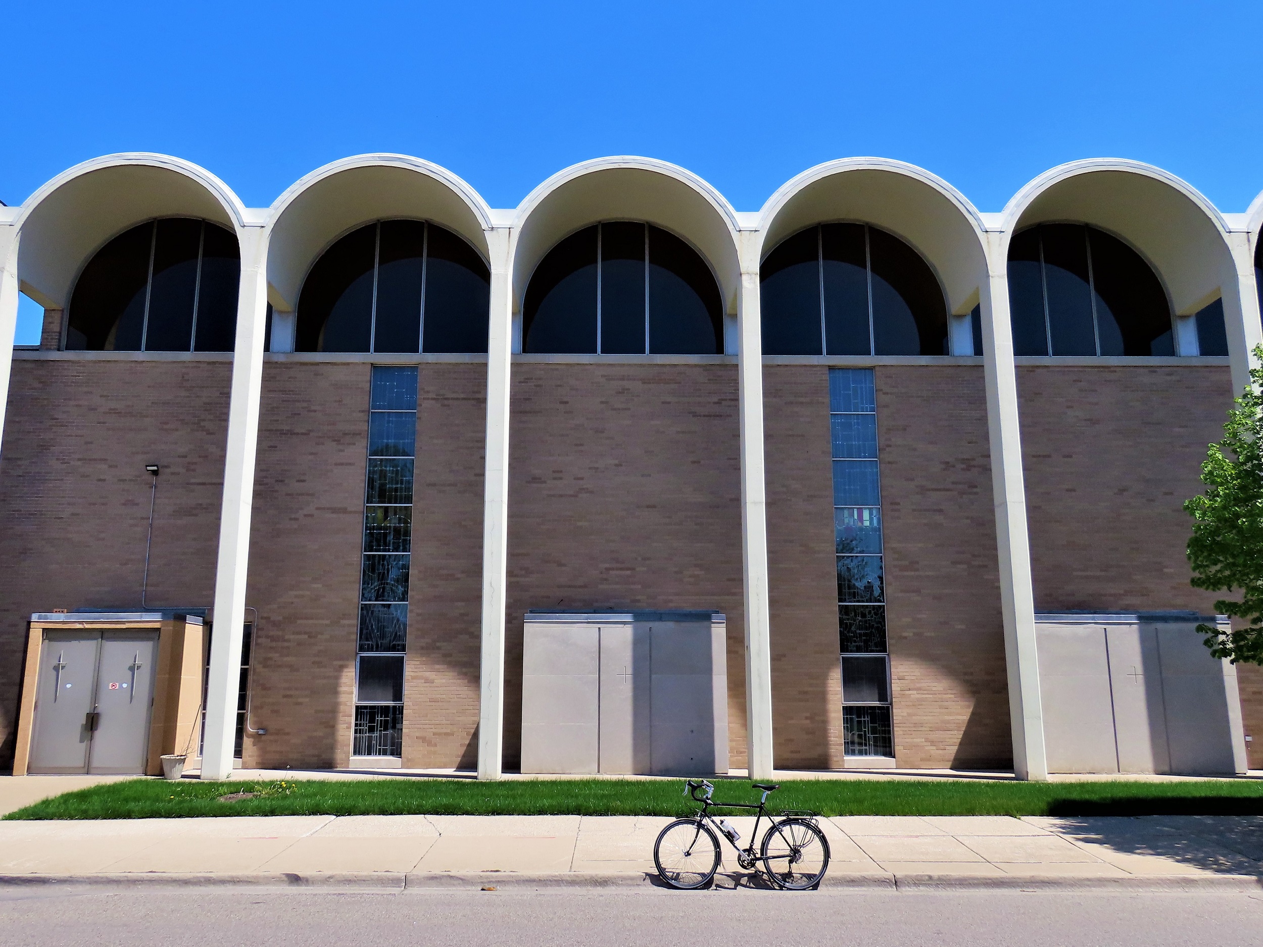 A tour bike standing at front of a series of white Mid Century Modern arches over a row of windows topping brown brick, three double doors are at the base.