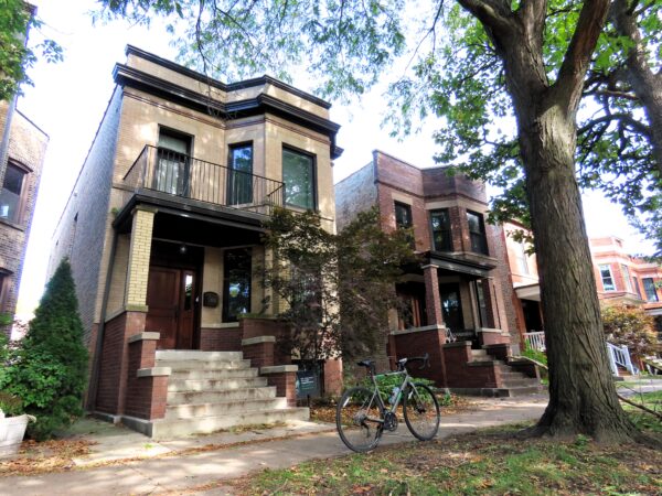 A tour bicycle standing in front of a yellow brick Chicago two flat with a black cornice under a canopy of trees.