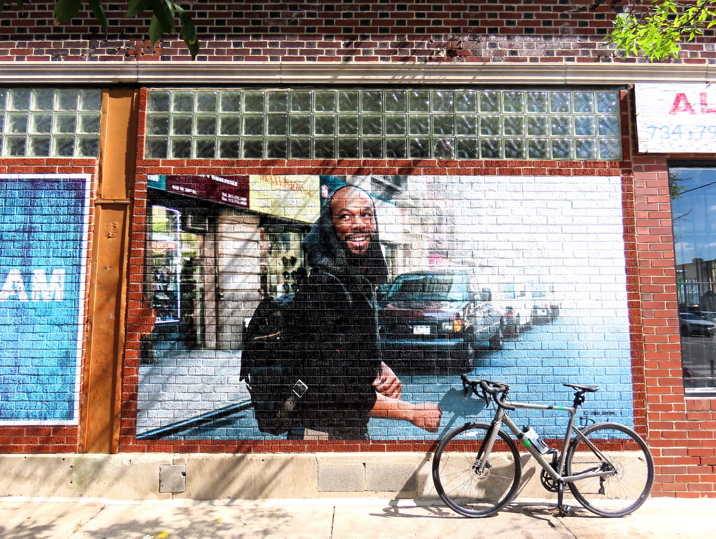 A tour bike leaning on a photorealistic mural of a smiling young Black man in a black hoody walking on the street.