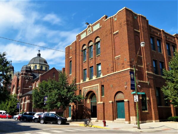 A tour bicycle standing in front of a red brick and Prairie Style tile accented three story school hall with a church dome in the distance.