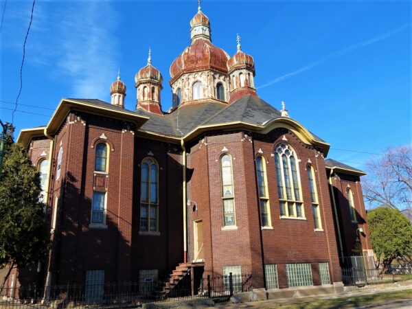 Seen from behind and to the side, a brown brick Byzantine style church with four visible rusted domes.