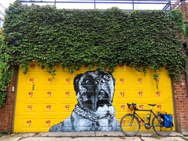 A tour bicycle leaning on a ivy topped garage painted yellow with a black and white mastiff dog and slices of pizza falling like rain.