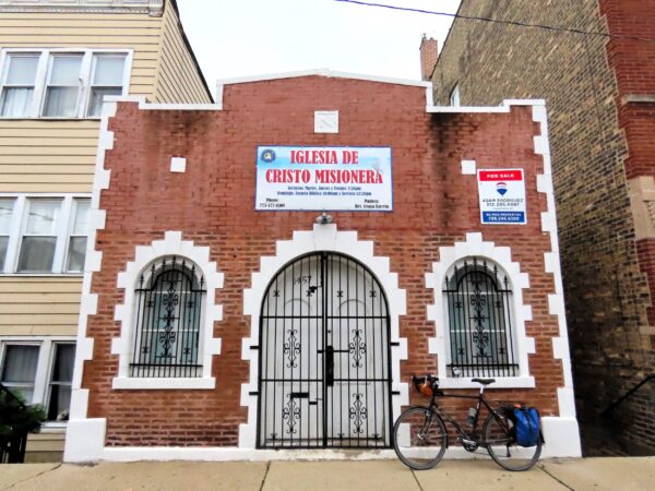 A tour bicycle leaning to the right of the entry of a one story red brick with white outline accents, Mission style reference.