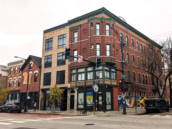 A tour bicycle standing at front of a red brick corner four story storefront and flats with a third/fourth floor corner bay, patina cornice and tan brick design accents.