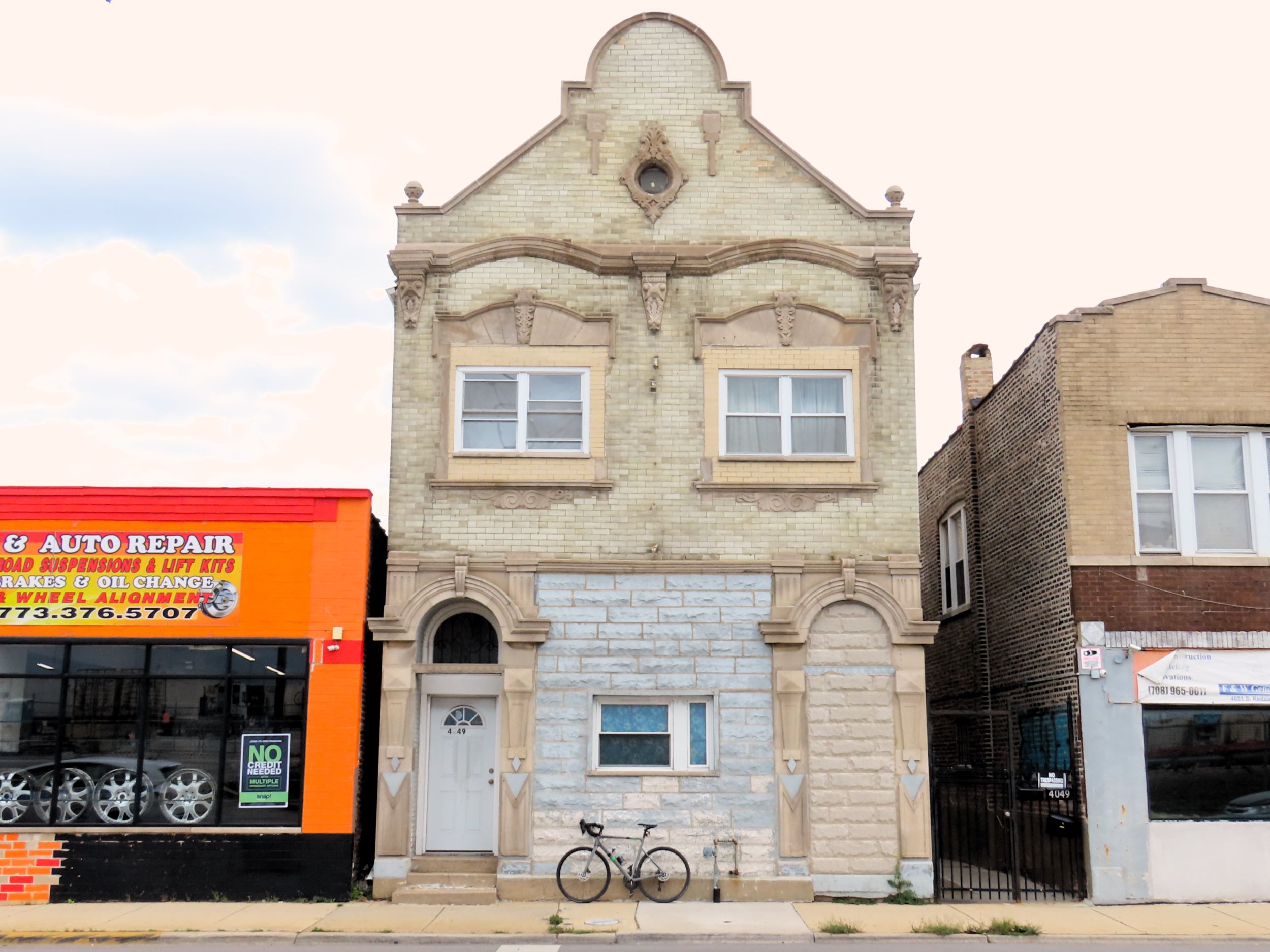 A tour bicycle leaning on the bottom of a pale brick three story storefront and flats with the storefront bricked up with grey block.