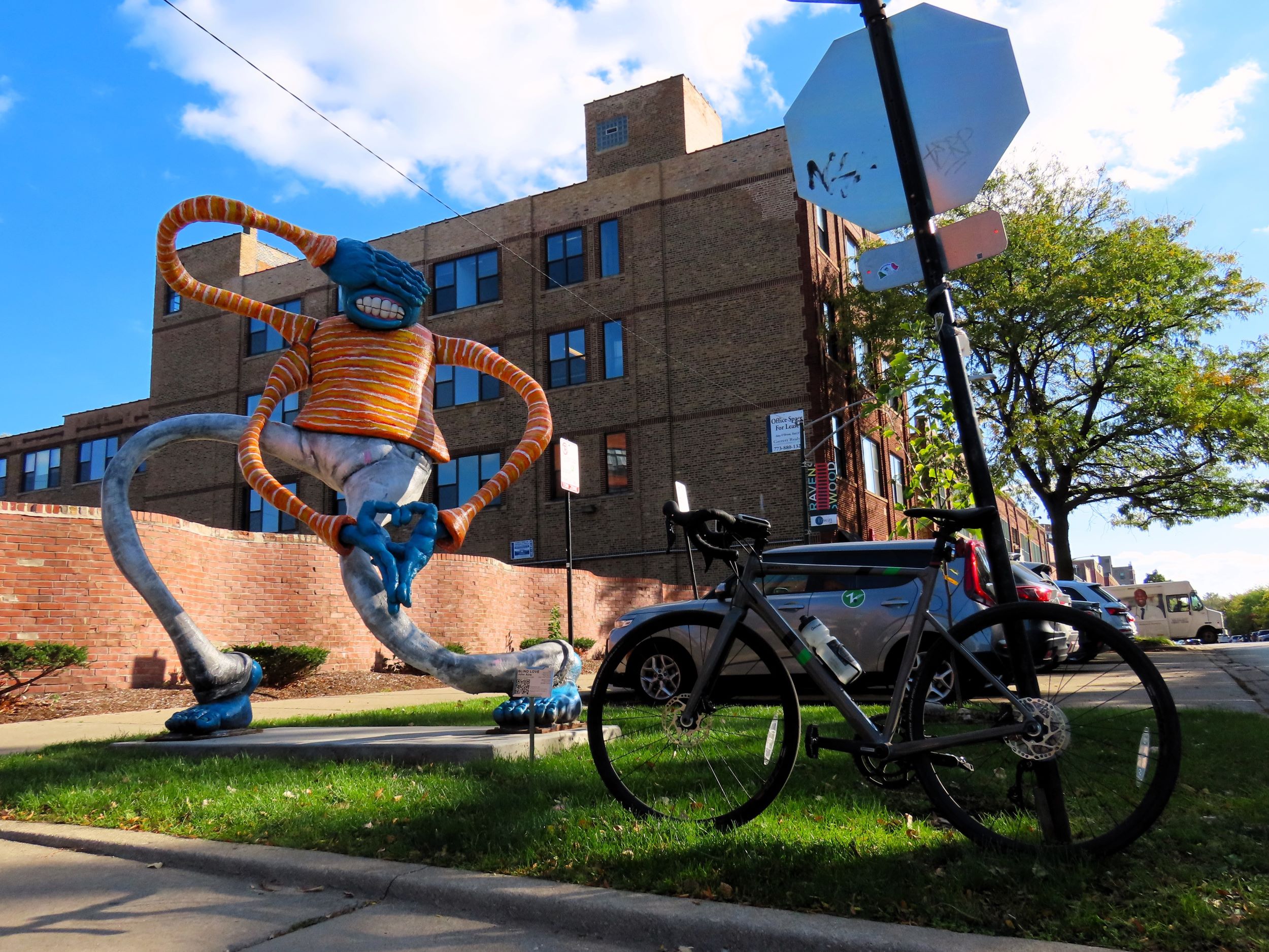 A tour bicycle standing to the side of a sculpture of a lanky blue man in a range sweater with three arms and one had covering his eyes.