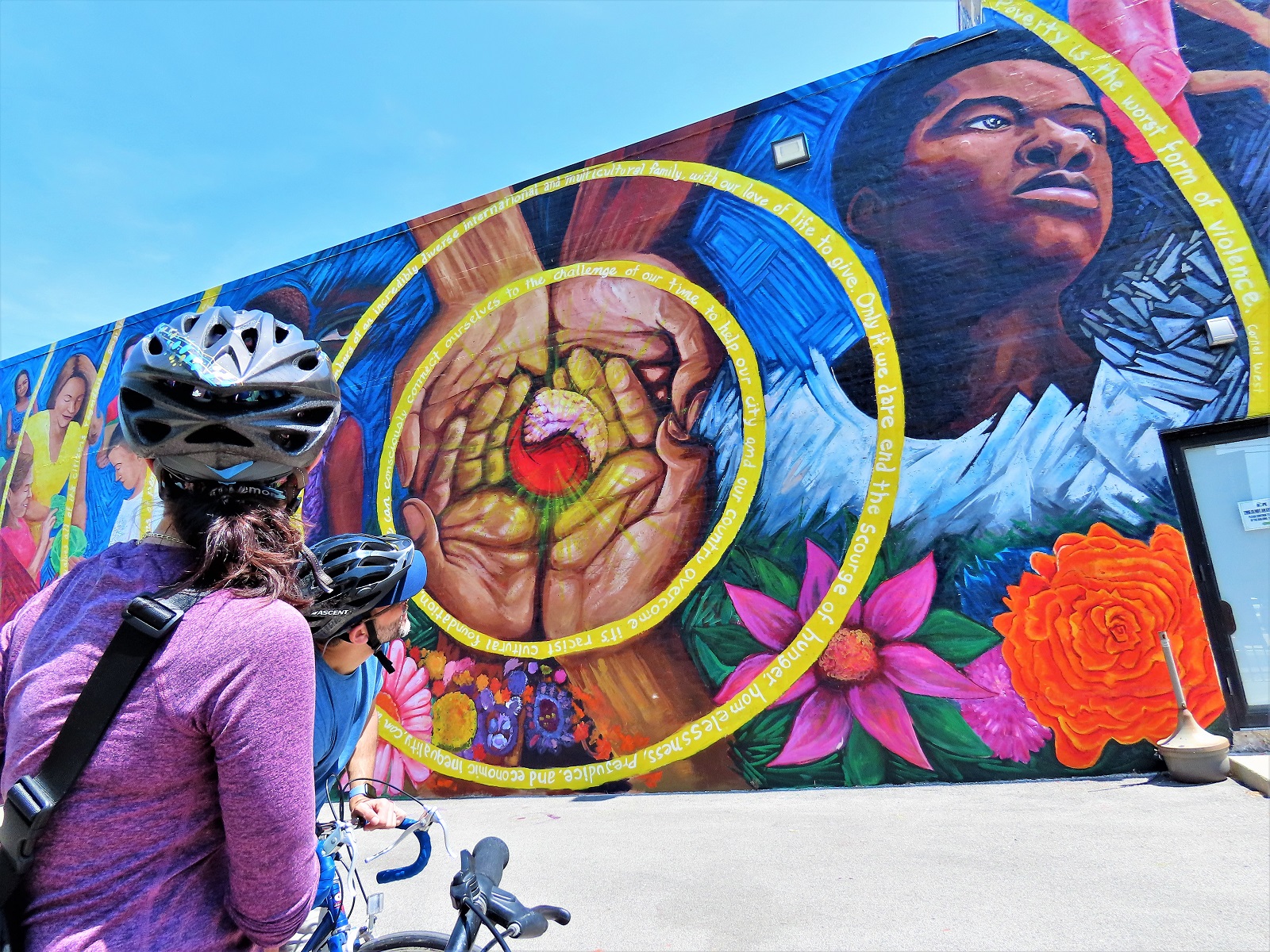Two CBA bike tour riders looking at a Mexican style mural with four hands holding a heart circled by two concentric yellow rings and a Black boy looking into the distance.