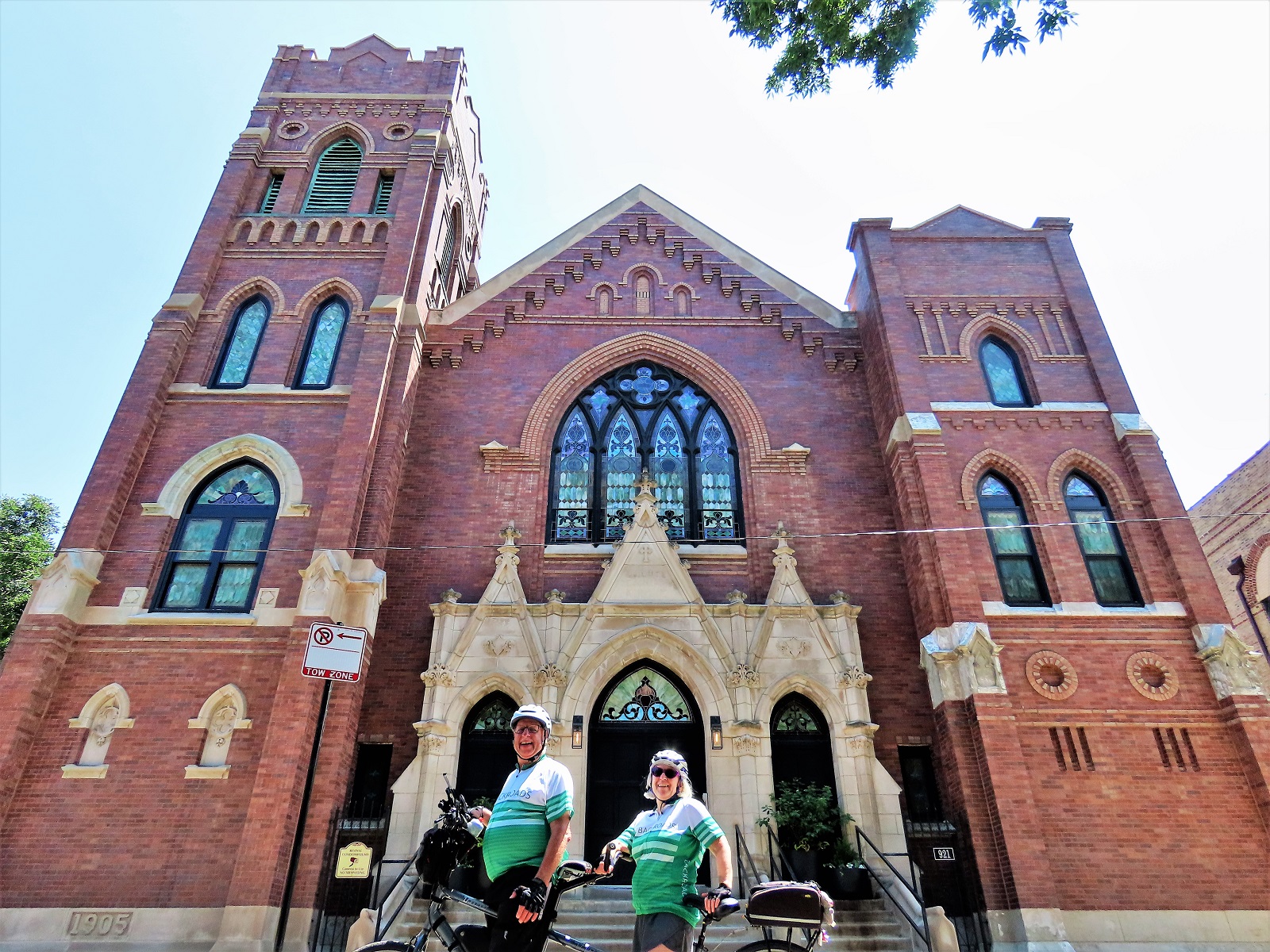 Two CBA friders on a tandem bike smiling at the camera in front of a brown brick Gothic Revival church with limestone entry.