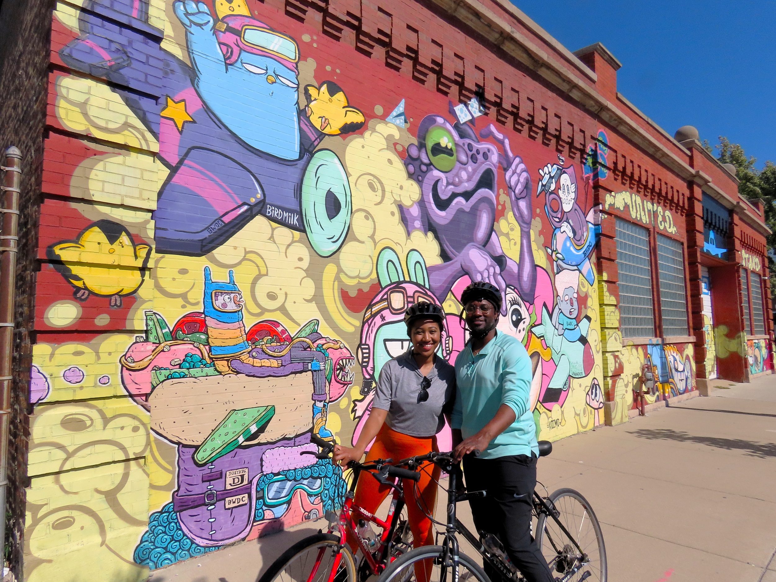 Two CBA riders arm in arm in front of colorful cartoon style street art on a red brick wall.