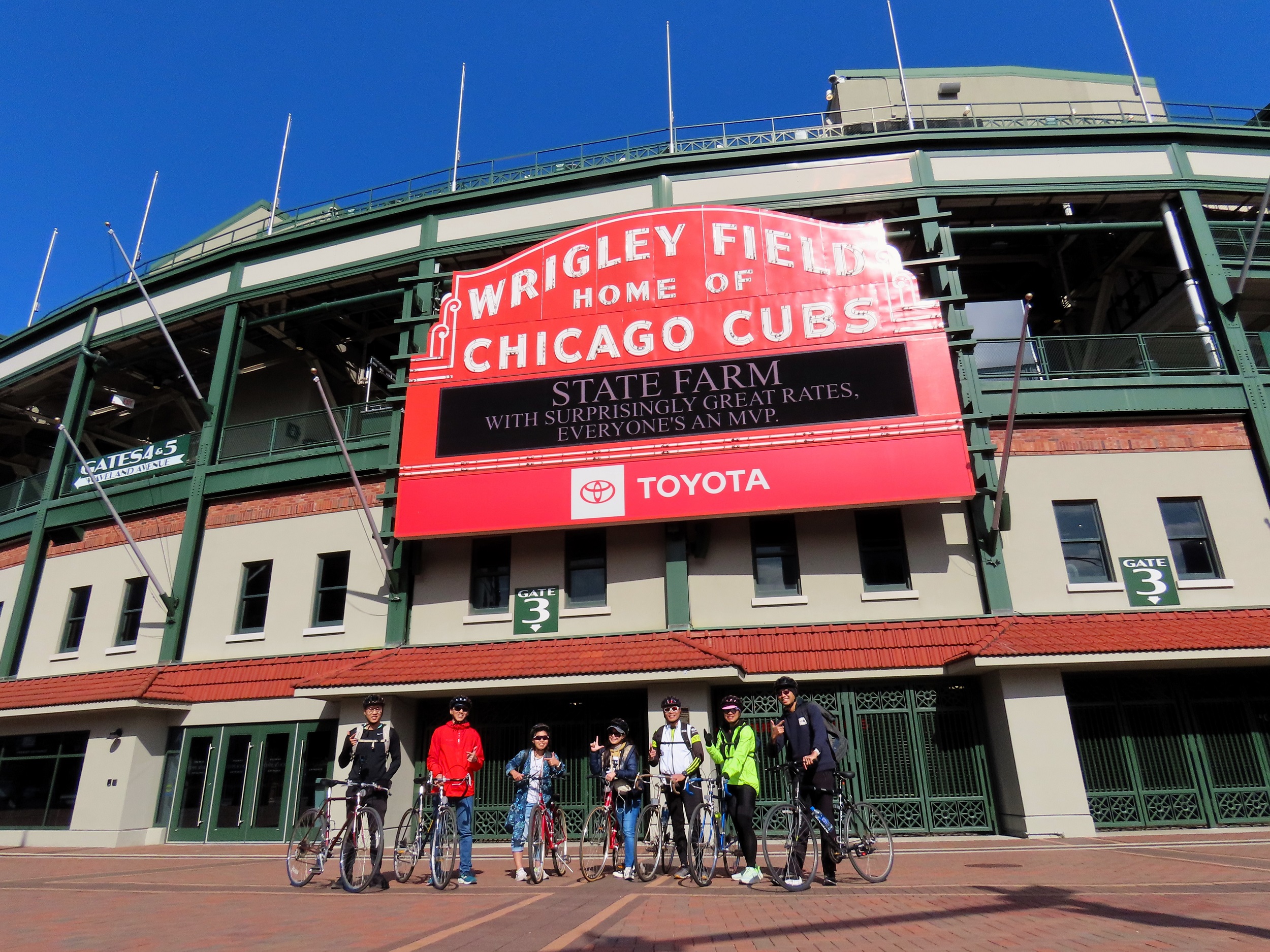 Seven CBA bike tour riders standing with thumbs up underneath the red Wrigley Field sign.