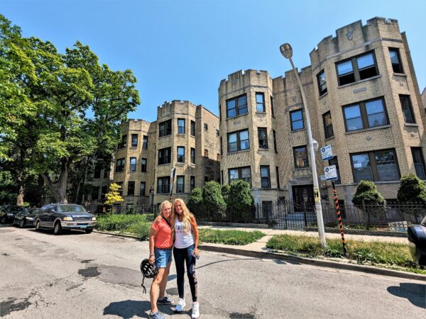 Two CBA riders smiling in front of a yellow brick three story 1920s apartment building.