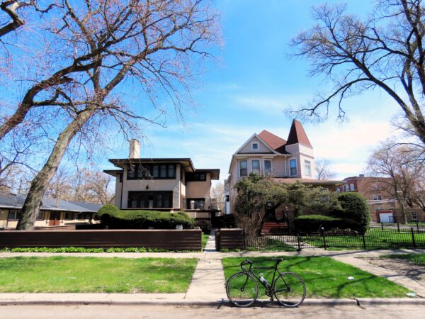 A tour bicycle is standing at front of two homes, on the left a two story earth tone Prairie School, the right a late 1800s pink wood Queen Anne.