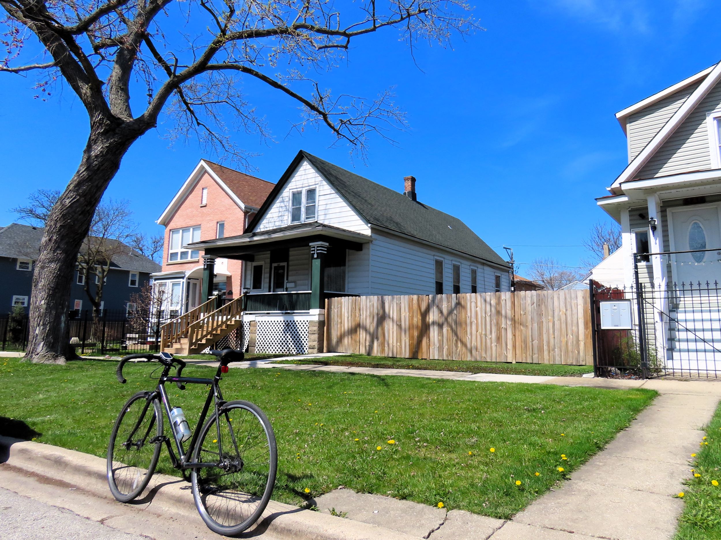 A tour bicycle standing in front of an early 1900s one and half story raised wood single family home with a large fenced side yard.