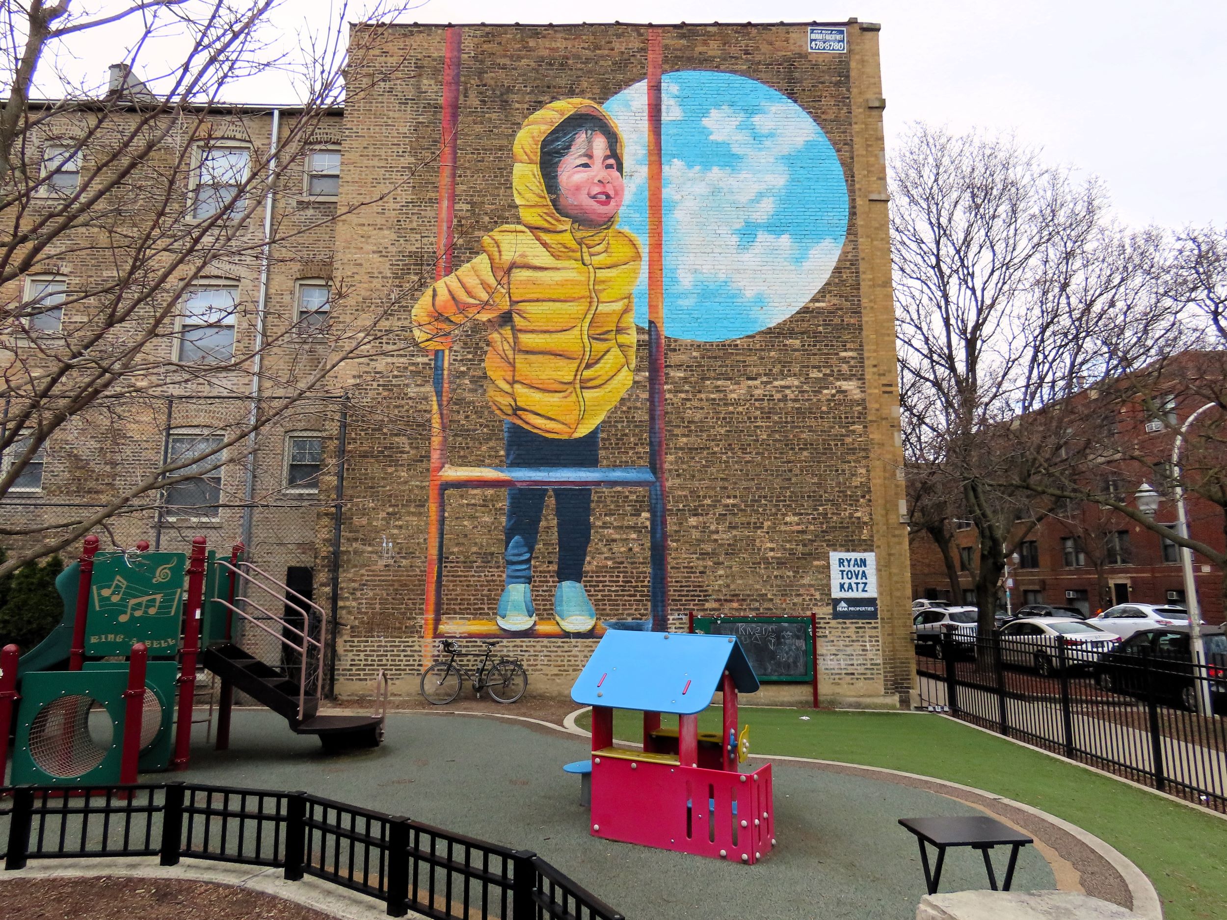 In a playground a tour bicycle is leaning on a brick wall below a three story high mural of a back haired child in a yellow jacket playing on a swing in front of the Earth.