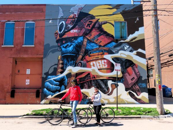 Two CBA riders standing with bikes in front of a two story mural of a cybernetic gorilla samurai.