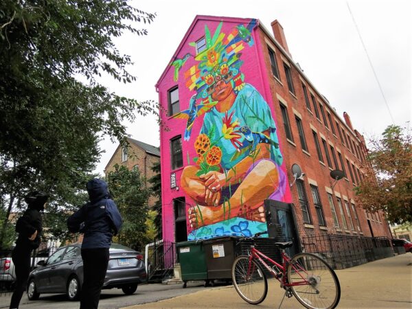 Two CBA riders standing in silhouette with bikes looking at a pink background mural with a Latin American indigenous person wearing an Aztec mask.