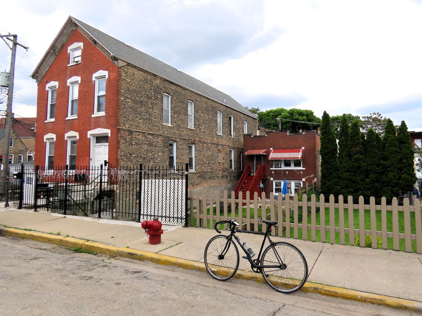 A tour bicycle standing in front of a row of two early 1900s brick buildings, on the left a three and half story multi unit, on the right a one story shoe box at the back of the lot with a visible far below street grade yard.