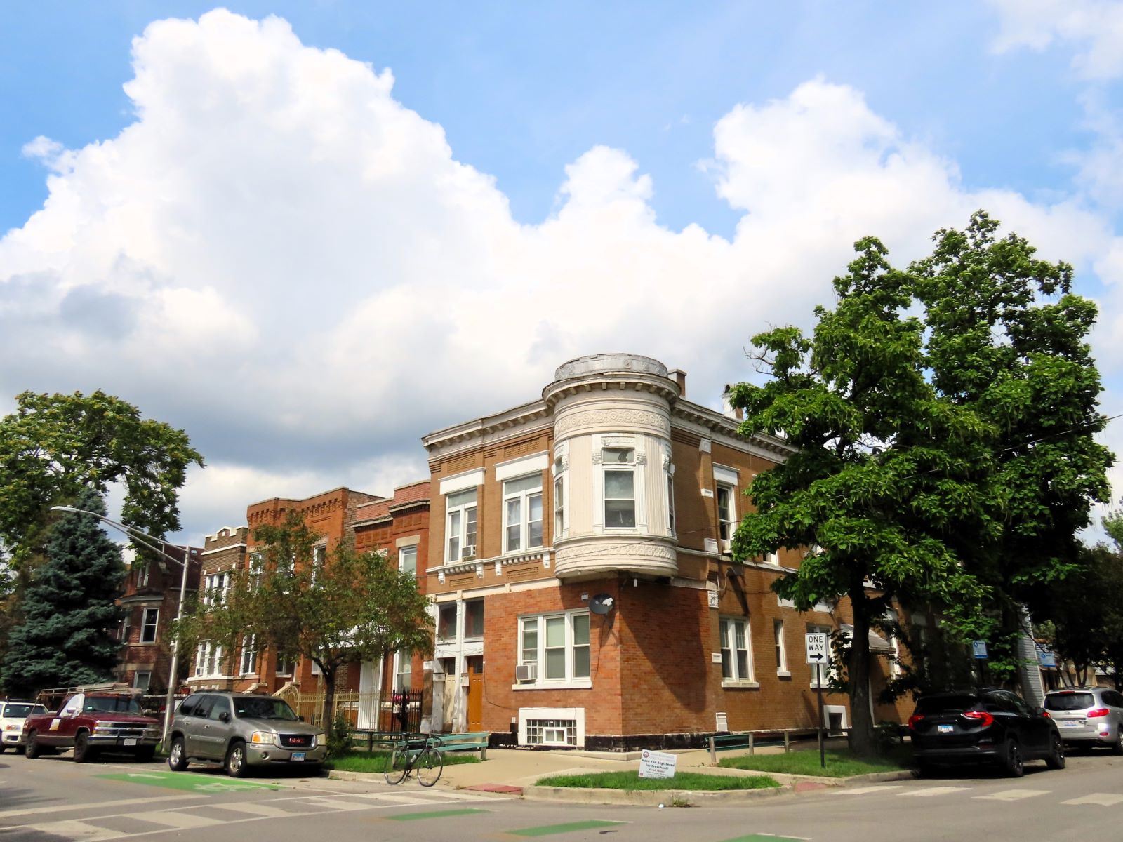 On a neighborhood street a tour bicycle is standing at front of a late 1800s second story turreted two story corner storefront and flats.