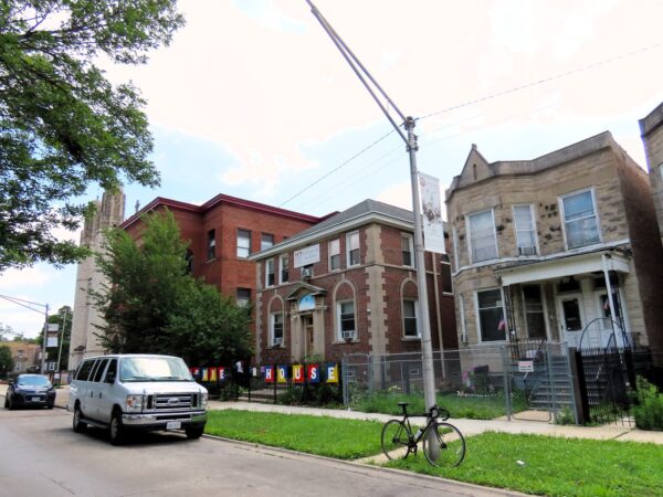 On a neighborhood street a tour bicycle is standing at front of an early 1900s greystone two flat with neighboring church building and Midcentury church towers in the distance.