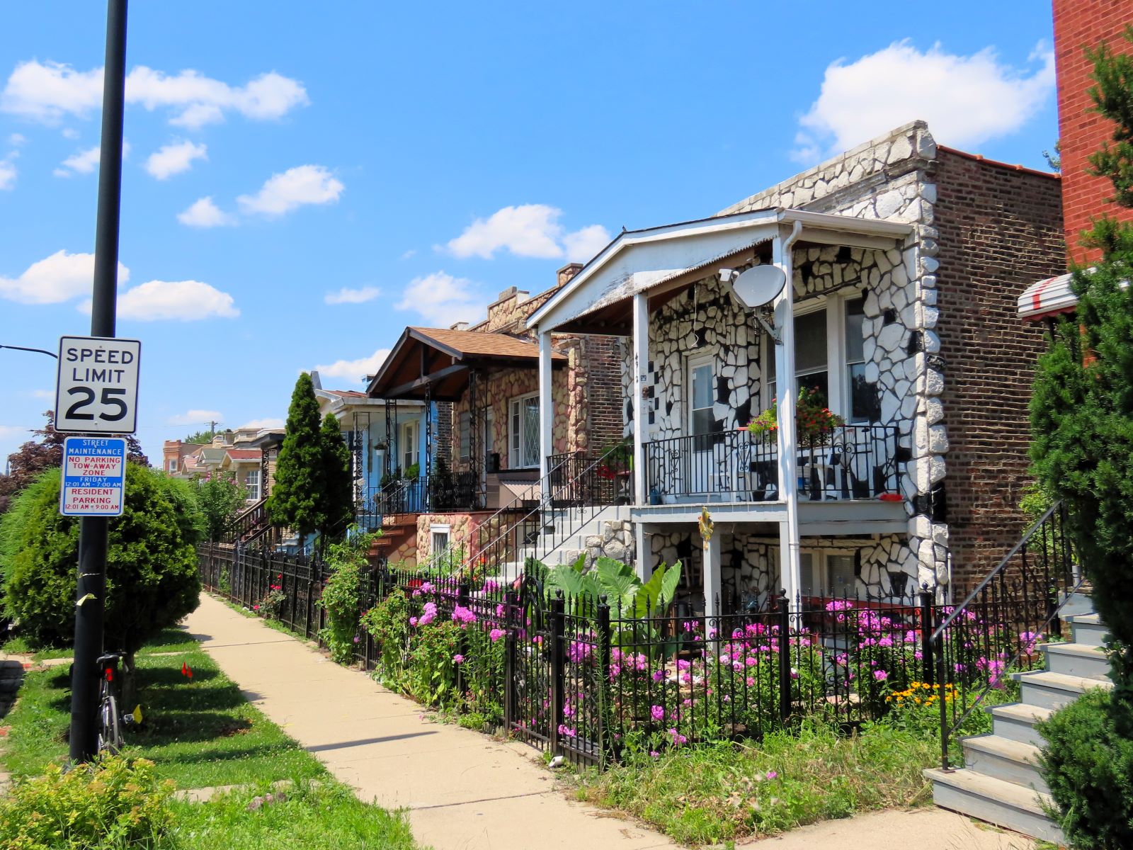 On a neighborhood street a tour bicycle is standing at front of three early 1900s raised one story flat roof homes with front porches, nearest with black and white permastone, next to that pink and white.