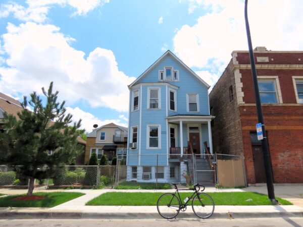 On a neighborhood street a tour bicycle is standing at front of an early 1900s blue sided wood frame pitched roof two and half story two flat.