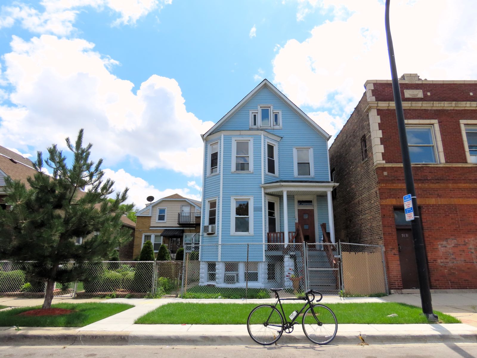 On a neighborhood street a tour bicycle is standing at front of an early 1900s blue sided wood frame pitched roof two and half story two flat.