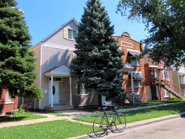 On a neighborhood street a tour bicycle is standing at front of an early 1900s sided wood frame two story home with a skinny pine in front.